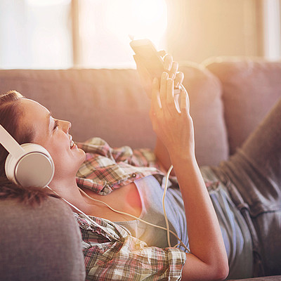 Eine Frau liegt auf dem Couch. Sie trägt Kopfhörer und schaut lächelnd auf dem Smartphone.